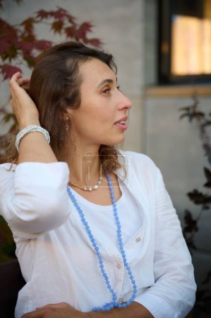 Téléchargez les photos : Portrait d'une jeune femme en chemise blanche et un collier autour de son cou par une journée ensoleillée dans un café avec de la verdure en arrière-plan - en image libre de droit