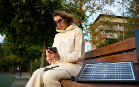 Photo for Young pretty woman charging her phone with a portable solar panel. Solar battery charger for mobile devices. Concept of sustainable lifestyle and green renewable energy - Royalty Free Image