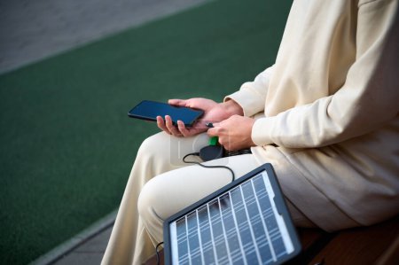 Téléchargez les photos : Jolie femme assise sur le banc et rechargeant son téléphone avec un panneau solaire portable. Chargeur de batterie solaire pour appareils mobiles. Concept de mode de vie durable et d'énergies renouvelables vertes. Vue des cultures - en image libre de droit