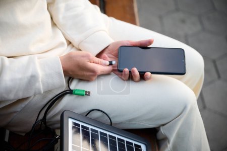 Photo for Pretty woman sitting on the bench and charging her phone with a portable solar panel. Solar battery charger for mobile devices. Concept of sustainable lifestyle and green renewable energy. Crop view - Royalty Free Image