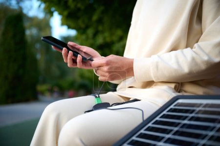 Foto de Mujer bonita sentada en el banco y cargando su teléfono con un panel solar portátil. Cargador de batería solar para dispositivos móviles. Concepto de estilo de vida sostenible y energía verde renovable. Vista de los cultivos - Imagen libre de derechos