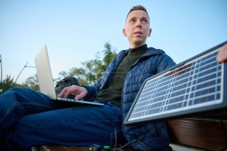Photo for Confident businessman sitting on the bench and charging his laptop with portable solar panel. Concept of renewable energy. Green independent energy. - Royalty Free Image
