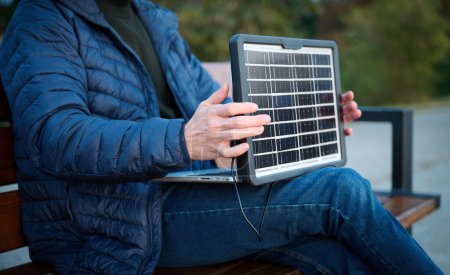 Téléchargez les photos : Homme d'affaires confiant assis sur le banc et chargeant son ordinateur portable avec un panneau solaire portable. Concept d'énergie renouvelable. Énergie verte indépendante. - en image libre de droit
