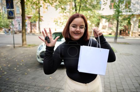 Photo for Portrait of young businesswoman shopping remotely using smartphone. Pretty female holding white paper bag with copy space, staying near the modern small car. Woman with car key. Rent car. - Royalty Free Image