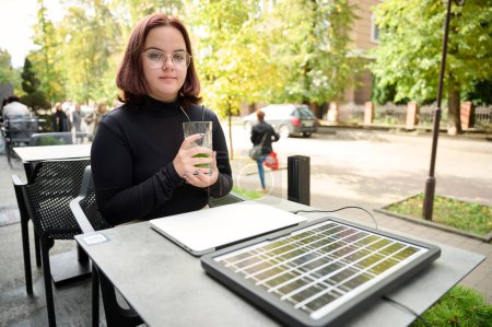 Téléchargez les photos : Femme utilisant un panneau solaire portable pour charger un ordinateur portable et un smartphone. Portrait d'une jeune étudiante travaillant à distance à partir d'un ordinateur portable alors qu'elle était assise à une table dans un café. Boire du mojito. Energies renouvelables. - en image libre de droit