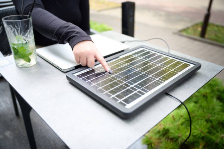 Photo for Female using portable solar panel to charge laptop and smartphone. Portrait of a young student girl working remotely from laptop while sitting at a table in a cafe. Pointing on solar cell. - Royalty Free Image