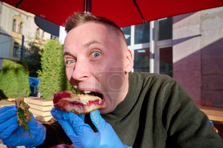 Téléchargez les photos : Homme caucasien affamé et drôle aime manger, déguster la délicieuse viande de hamburger avec du fromage et des légumes dans le café assis près de l'ordinateur portable. Restauration rapide malsaine. - en image libre de droit