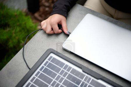 Foto de Vista de cerca de una chica conectando un enchufe USB a su computadora portátil para cargar desde un panel solar portátil. Hembra usando el panel solar portátil para cargar el ordenador portátil y el teléfono inteligente. Energía renovable. Imagen recortada - Imagen libre de derechos