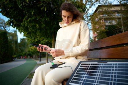 Téléchargez les photos : Jeune jolie femme chargeant son téléphone avec un panneau solaire portable. Chargeur de batterie solaire pour appareils mobiles. Concept de mode de vie durable et d'énergies renouvelables vertes - en image libre de droit
