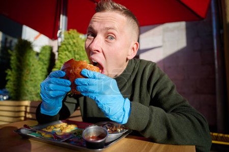 Téléchargez les photos : Homme caucasien affamé et drôle aime manger, déguster la délicieuse viande de hamburger avec du fromage et des légumes dans le café assis près de l'ordinateur portable. Restauration rapide malsaine. - en image libre de droit