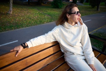 Foto de Retrato de una joven con gafas de sol modernas sentada en el banco de madera en un parque cerca de un lago disfrutando de la puesta de sol.Elegante chica con ropa deportiva ligera sentada y relajada en la playa al atardecer - Imagen libre de derechos