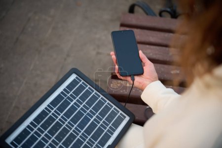 Foto de Mujer bonita joven cargando su teléfono con un panel solar portátil. Cargador de batería solar para dispositivos móviles. Concepto de estilo de vida sostenible y energía verde renovable - Imagen libre de derechos