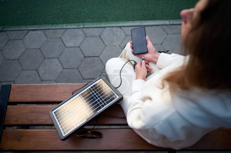 Foto de Mujer bonita joven cargando su teléfono con un panel solar portátil. Cargador de batería solar para dispositivos móviles. Concepto de estilo de vida sostenible y energía verde renovable - Imagen libre de derechos