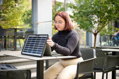 Photo for Female using portable solar panel to charge laptop and smartphone. Portrait of a young student girl working remotely from laptop while sitting at a table in a cafe. Bright sunny day. Renewable energy. - Royalty Free Image
