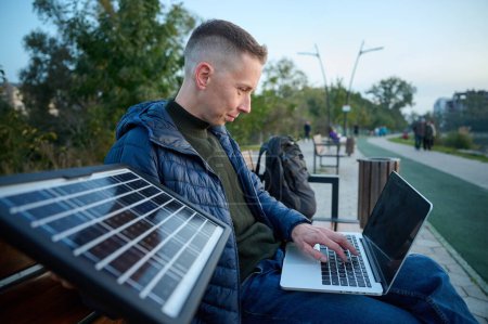 Téléchargez les photos : Homme d'affaires confiant assis sur le banc et chargeant son ordinateur portable avec un panneau solaire portable. Concept d'énergie renouvelable. Énergie verte indépendante. Vue latérale - en image libre de droit