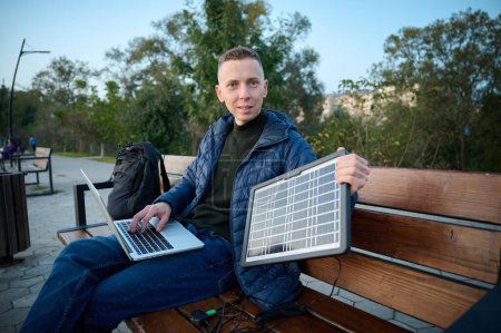 Photo for Confident businessman sitting on the bench and charging his laptop with portable solar panel. Concept of renewable energy. Green independent energy. - Royalty Free Image