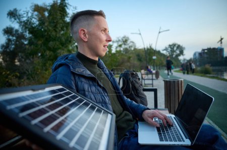 Téléchargez les photos : Homme d'affaires confiant assis sur le banc et chargeant son ordinateur portable avec un panneau solaire portable. Concept d'énergie renouvelable. Énergie verte indépendante. Vue latérale - en image libre de droit