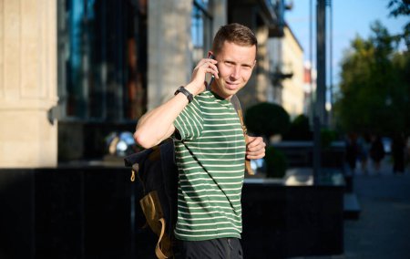 Photo for Young sporty man in a striped T-shirt walking on the street with a smartphone in hand and a trendy hipster's backpack slung over his shoulder, enjoying a leisurely urban outing. - Royalty Free Image