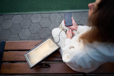 Téléchargez les photos : Jeune jolie femme chargeant son téléphone avec un panneau solaire portable. Chargeur de batterie solaire pour appareils mobiles. Concept de mode de vie durable et d'énergies renouvelables vertes - en image libre de droit