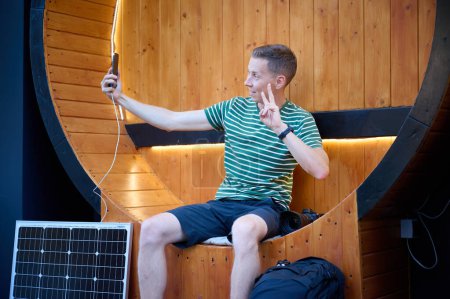 Photo for Excited photographer looks at the photos he just took. Young photographer sitting and checking result of photosession. Handsome male sitting on round bench with solar panel on background. - Royalty Free Image