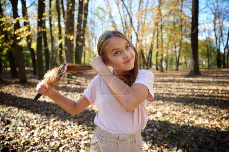 Foto de Retrato de una joven adolescente peinándose el pelo con un peine. Brillante día soleado de otoño en el parque con hojas caídas. Preparación para la sesión de fotos. - Imagen libre de derechos