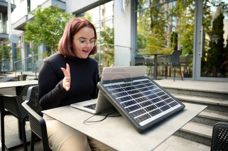Photo for Female using portable solar panel to charge laptop and smartphone. Portrait of a young student girl working remotely from laptop while sitting at a table in a cafe. Surprised woman. Renewable energy. - Royalty Free Image