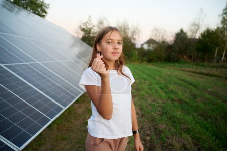 Téléchargez les photos : Jolie jeune fille restant près des panneaux solaires et montrant le symbole du cœur. Concept d'énergie solaire. Les énergies renouvelables. Panneaux photovoltaïques - en image libre de droit