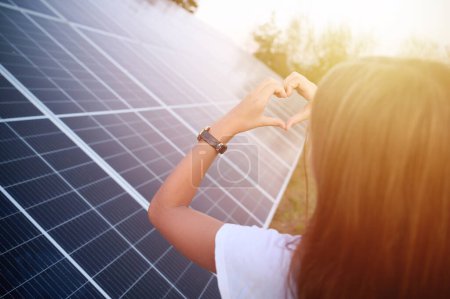 Foto de Linda joven que se queda cerca de paneles solares y muestra el símbolo del corazón. Concepto de energía solar. Energía renovable. Vista trasera. - Imagen libre de derechos