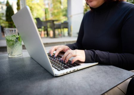 Photo for Cropped image of the student girl typing on laptop while sitting at a table in a cafe. Female student studying at park using modern laptop at bright sunny day. Working outdoor. Side view. - Royalty Free Image