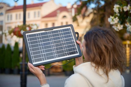Photo for Young pretty woman charging her phone with a portable solar panel. Solar battery charger for mobile devices. Concept of sustainable lifestyle and green renewable energy - Royalty Free Image
