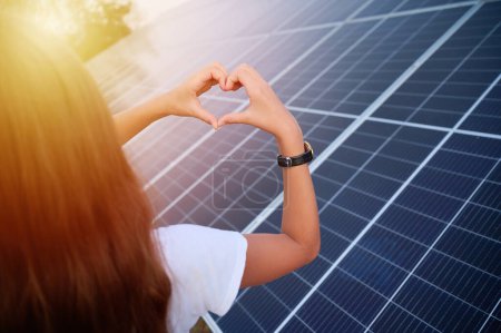 Photo for Pretty young girl staying near solar panels and showing heart symbol. Solar energy concept. Renewable energy. Back view. - Royalty Free Image