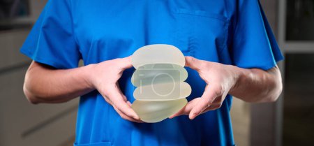 Foto de El primer plano de la cirujana plástica femenina muestra los implantes mamarios a una paciente para su nueva mama. El cirujano plástico sostiene los implantes de silicona mamaria. Concepto de aumento de senos. Muestras de implantes mamarios - Imagen libre de derechos
