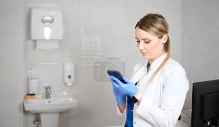 Pretty female doctor in blue medical gloves holding smartphone using mobile healthcare tech app consulting remote patient online. Gynecologist woman in her gynecological office.