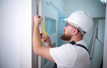 Photo for Construction worker with a tape in hands fixing at construction site. Accessories for assembling, install furniture, repair home. Man dressed in work attire, helmet and protective glasses. Home renovation concept - Royalty Free Image