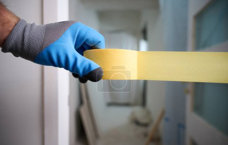 Photo for Worker with a masking tape in hands at construction site. Quick and easy painting process. Accessories for install furniture, repair home. Man dressed in blue gloves. Home renovation concept - Royalty Free Image