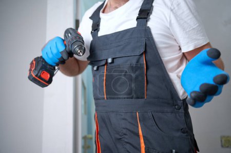 Photo for Construction worker holding electric screwdriver in hand at construction site. Accessories for assembling, install furniture, repair home. Man dressed in work attire and helmet and protective glasses. - Royalty Free Image