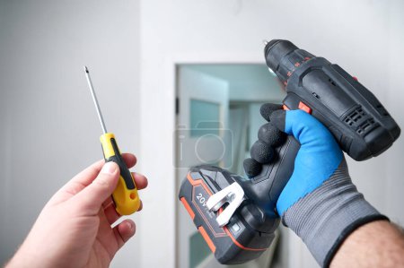 Photo for Worker holding electric screwdriver and simple screwdriver in hand at construction site. Accessories for assembling, install furniture, repair home. Home renovation, electric tool for job. Crop view. - Royalty Free Image