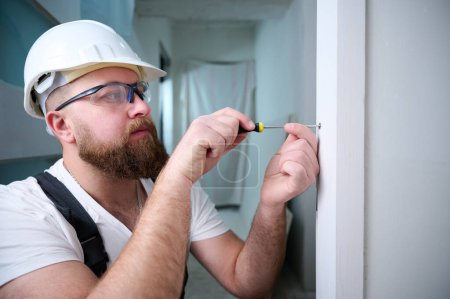 Photo for Construction worker with a screwdriver installing a door. Accessories for assembling, install furniture, repair home. Man dressed in work attire, helmet and protective glasses. Home renovation concept - Royalty Free Image