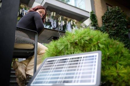 Frauen nutzen tragbare Solarzellen, um Laptop und Smartphone aufzuladen. Porträt einer jungen Studentin, die ferngesteuert vom Laptop an einem Tisch in einem Café sitzt. Erneuerbare Energien. Geringer Kamerawinkel