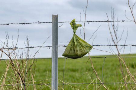 Un primer plano de una bolsa de caca de perro colgando de una valla en el campo