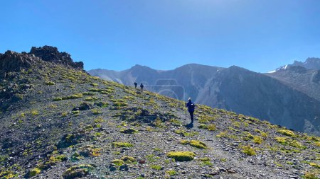 Foto de Un grupo de turistas está caminando cuesta arriba. Parque Nacional Ala-Archa, Kirguistán. Vista de las colinas de montaña, rocas y laderas. Hermoso paisaje de verano de montaña. Fondo natural tranquilo. - Imagen libre de derechos