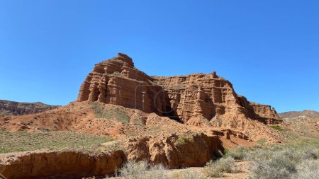 The canyons of Konorchek. Steep rocky geological formations in Kyrgyzstan. A red rocky mountain with a clear blue cloudless sky in the background. A grassy area.