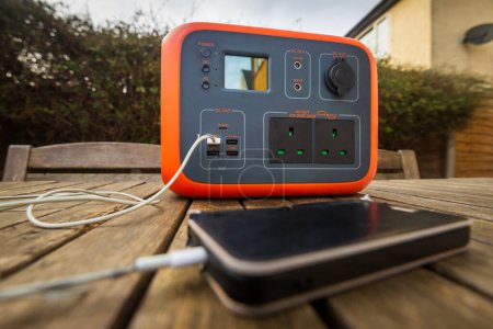 Portable power station solar electricity generator outdoors with mobile phone plugged in to charge. Wireless charging lithium battery backup for power outage, emergencies, travel or camping.