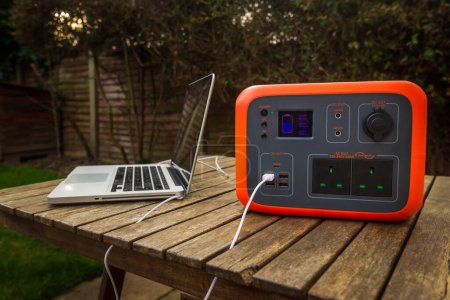 Portable power station solar electricity generator outdoors with laptop plugged in charging. Wireless charging lithium battery backup for power outage emergencies outdoors, camping or travel.