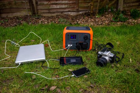 Portable power station solar electricity generator with laptop, tablet and camera electronic devices charging outdoors on garden lawn grass. Wireless charging lithium battery backup for use anywhere.