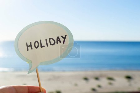 Photo for Stick with paper speech bubble with words Holiday on background blue sea, sky, sandy beach on sunny summer day. Text-balloons with text from letters. Concept, symbol, sign vacation travel tourism rest - Royalty Free Image