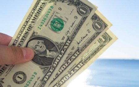 Téléchargez les photos : Homme tenant trois billets d'un dollar dans sa main sur fond de mer bleue et de ciel bleu le jour ensoleillé de l'été. Le concept d'argent, finance, monétaire, voyage, tourisme, vacances, repos - en image libre de droit