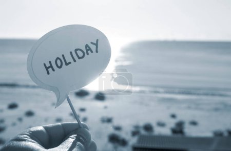 Photo for Stick with paper speech bubble with words Holiday on background blue sea, sky, sandy beach on sunny summer day. Text-balloons with text from letters. Concept, symbol, sign vacation travel. Blue color - Royalty Free Image