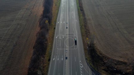 Photo for Cars driving along the highway on an autumn sunny day. Automobile road with white markings between agricultural fields. View of the cars driving on paved road. automobile vehicles. Aerial drone view. - Royalty Free Image