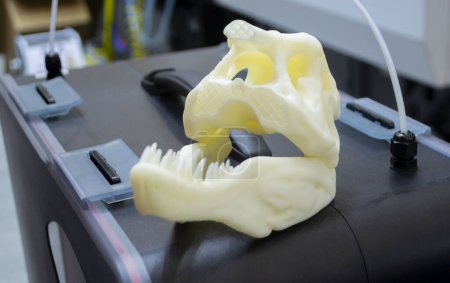 Foto de Objeto de arte impreso en impresora 3D. Prototipo de cabeza de cráneo de dinosaurio con dientes. Modelo impreso en impresora 3D de plástico fundido. Objeto impreso de cerca. Concepto nueva tecnología de impresión de innovación moderna - Imagen libre de derechos